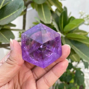May include: A purple amethyst crystal shaped like a hexagon with many facets. The crystal is being held in a hand.