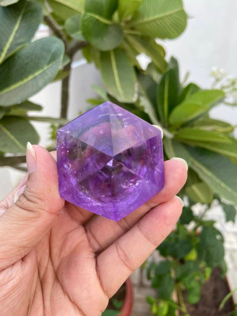 May include: A purple amethyst crystal shaped like a hexagon with many facets. The crystal is being held in a hand.