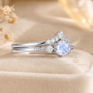May include: A silver-toned ring set featuring a round, iridescent moonstone centre stone surrounded by a halo of small, clear, round-cut stones. The band has a delicate, textured metal design with additional small stones embedded along the shank.