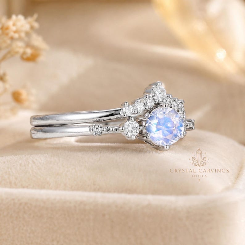 May include: A silver-toned ring set featuring a round, iridescent moonstone centre stone surrounded by a halo of small, clear, round-cut stones. The band has a delicate, textured metal design with additional small stones embedded along the shank.