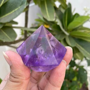 May include: A purple amethyst crystal pyramid with a smooth, polished surface. The pyramid is a geometric shape with a pointed top and a square base.