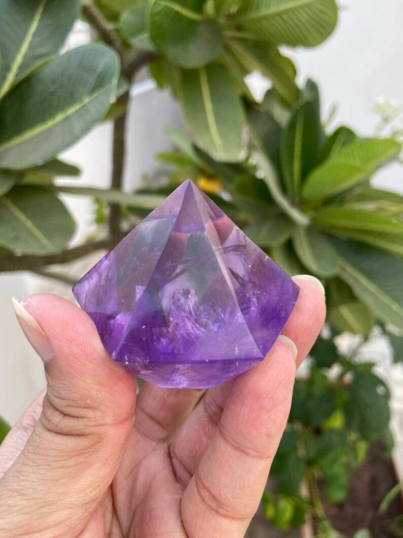 May include: A purple amethyst crystal pyramid with a smooth, polished surface. The pyramid is a geometric shape with a pointed top and a square base.
