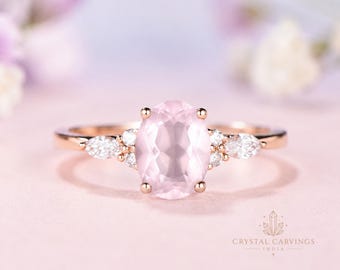 May include: A rose gold ring featuring a central oval-cut pale pink gemstone, flanked by two small marquise-cut clear stones on each side, accented with tiny round clear stones set in a prong setting on a polished rose gold band.