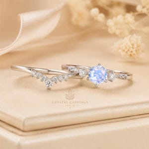 May include: A silver-toned ring set featuring a round, iridescent moonstone centre stone flanked by small clear accent stones on a thin band, paired with a matching chevron-shaped band adorned with a row of small clear stones and text.