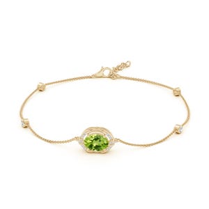 May include: A gold chain bracelet featuring a central oval-cut green peridot gemstone surrounded by small white diamonds. The delicate chain is accented with four evenly spaced round white diamonds set in gold bezels and a lobster claw clasp.