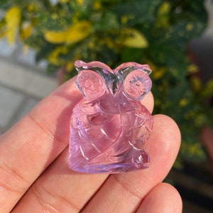 Puede incluir: Una figurita de búho tallada en amatista con un tono rosa claro. El búho está detallado con tallas intrincadas y tiene una apariencia ligeramente translúcida.