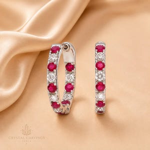 May include: A pair of silver hoop earrings featuring an alternating pattern of round-cut red rubies and clear white diamonds. The earrings have a hinged closure mechanism and are displayed upright, showcasing the vibrant red and sparkling white stones.