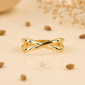 Peut inclure: Une bague en or poli présentant un design croisé lisse où deux anneaux s'entrecroisent au centre. La bague possède une finition métallique réfléchissante et une forme fluide continue qui crée une silhouette ouverte en forme de X sur le devant.