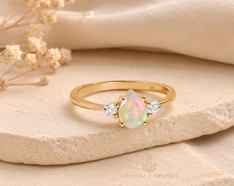 May include: A gold ring featuring a central pear-shaped opal flanked by two small round clear stones. The band is thin and polished, with a 925 hallmark visible on the inner surface. The design centres on the iridescent gemstone.