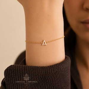 Puede incluir: Una delicada pulsera de cadena dorada con un colgante en forma de letra A incrustado con pequeñas piedras transparentes. La letra A está centrada en la fina cadena metálica que se sujeta alrededor de la muñeca. El diseño es sencillo y refinado.