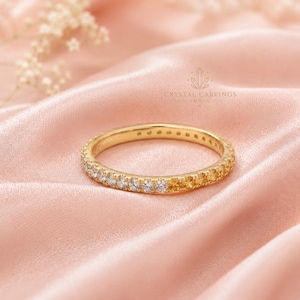 May include: A gold band ring featuring a continuous row of small, round-cut gemstones that transition in color from clear white to warm yellow. The ring is positioned on a soft, draped pink fabric. The text CRYSTAL CARVINGS INDIA is visible.