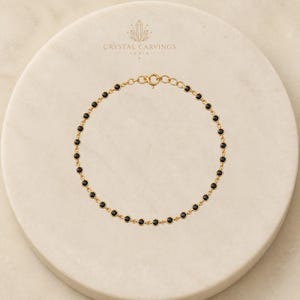 May include: A gold chain bracelet featuring a series of small, round, black stones set in individual gold bezels. The bracelet is arranged in a circular shape and includes a small gold spring ring clasp at the top. The text Crystal Carvings India is visible.
