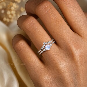 May include: A silver-toned ring set featuring a central round iridescent moonstone flanked by small clear stones, paired with a chevron-shaped band adorned with a row of tiny clear stones. The metal bands are thin and polished with a bright finish.