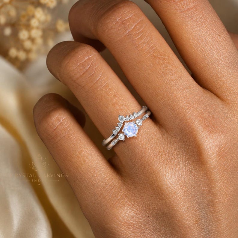 May include: A silver-toned ring set featuring a central round iridescent moonstone flanked by small clear stones, paired with a chevron-shaped band adorned with a row of tiny clear stones. The metal bands are thin and polished with a bright finish.