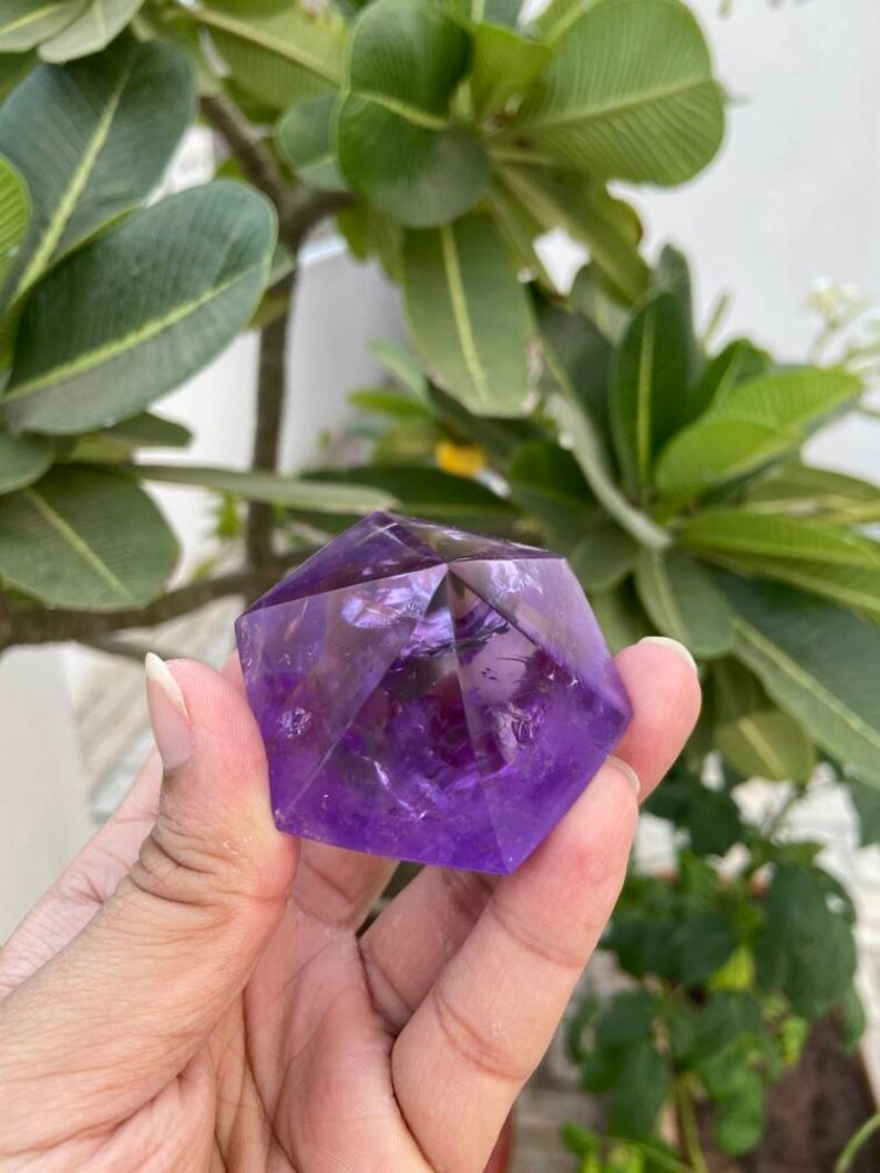 May include: A purple amethyst crystal carved into a geometric shape. The crystal is faceted and has a translucent appearance.