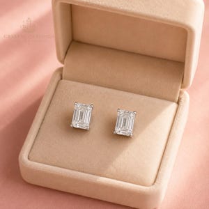 May include: A pair of emerald-cut stud earrings with clear, faceted stones set in silver-toned metal. The earrings are displayed inside a beige velvet jewellery box. The text CRYSTAL CARVINGS INDIA is printed in the upper left corner of the image.