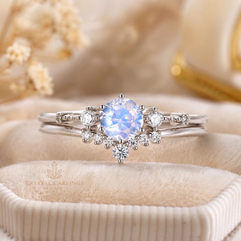 May include: A silver-toned ring set featuring a central round iridescent moonstone cabochon flanked by small clear round stones. The band includes a curved accent piece adorned with additional small clear stones arranged in a cluster design.