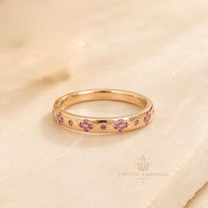 Puede incluir: Un anillo de banda de oro rosa con un patrón repetitivo de pequeñas piedras preciosas redondas de color rosa dispuestas en forma de flor. La superficie metálica es pulida y lisa, con el texto CRYSTAL CARVINGS INDIA en la esquina inferior derecha.