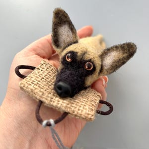 May include: A handmade felt dog toy with a tan and black color scheme. The dog has large, upright ears and is holding a small burlap sack with a brown handle. The toy is detailed with realistic features and is designed for pet play.