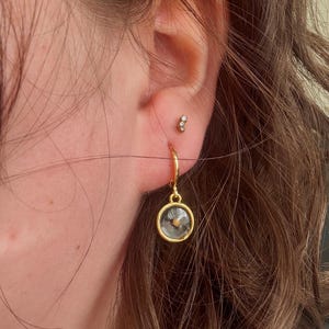 May include: Gold hoop earrings with a small, clear glass charm featuring a dried flower inside. The charm is hanging from the hoop.
