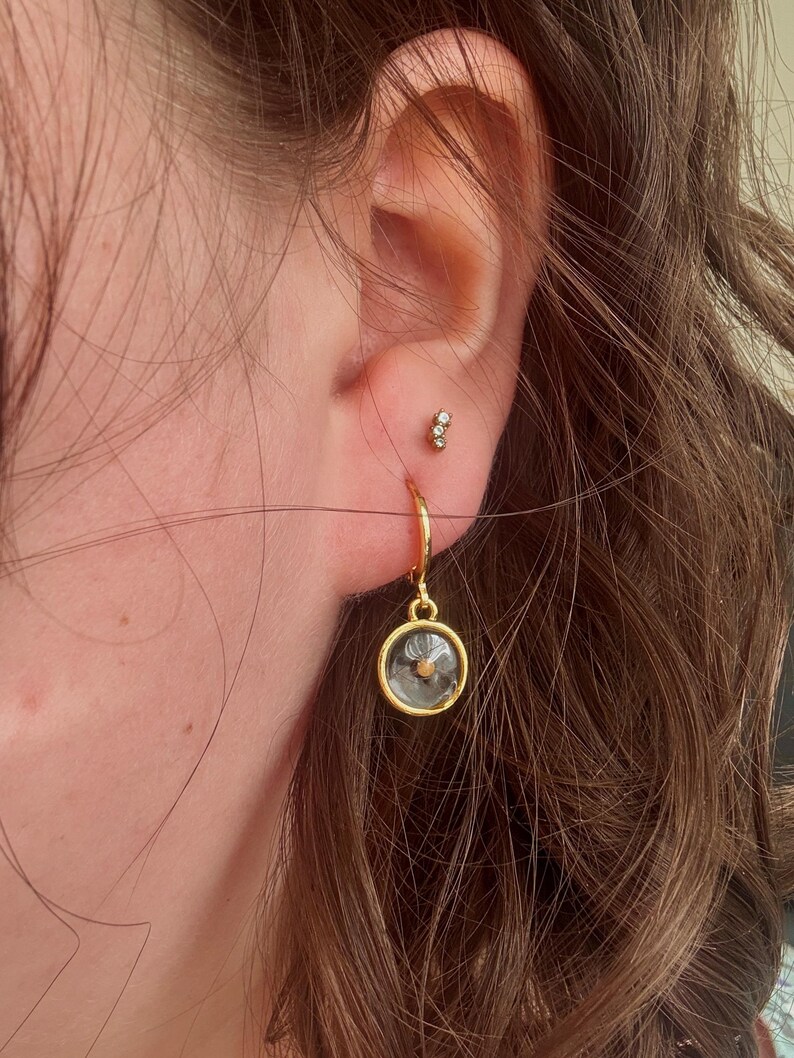 May include: Gold hoop earrings with a small, clear glass charm featuring a dried flower inside. The charm is hanging from the hoop.