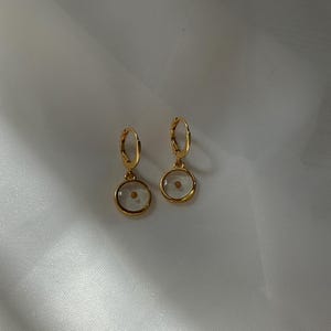 May include: A pair of gold hoop earrings with a small, clear circle pendant containing a single, tiny seed. The earrings are on a white background.