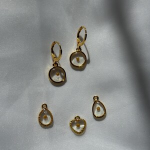 May include: Five gold-toned earrings and pendants with clear glass charms containing a single small seed. The charms are in the shapes of a circle, teardrop, and heart.