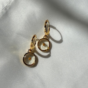 May include: A pair of gold hoop earrings with a small, clear circle pendant containing a single seed. The earrings are on a white fabric background.