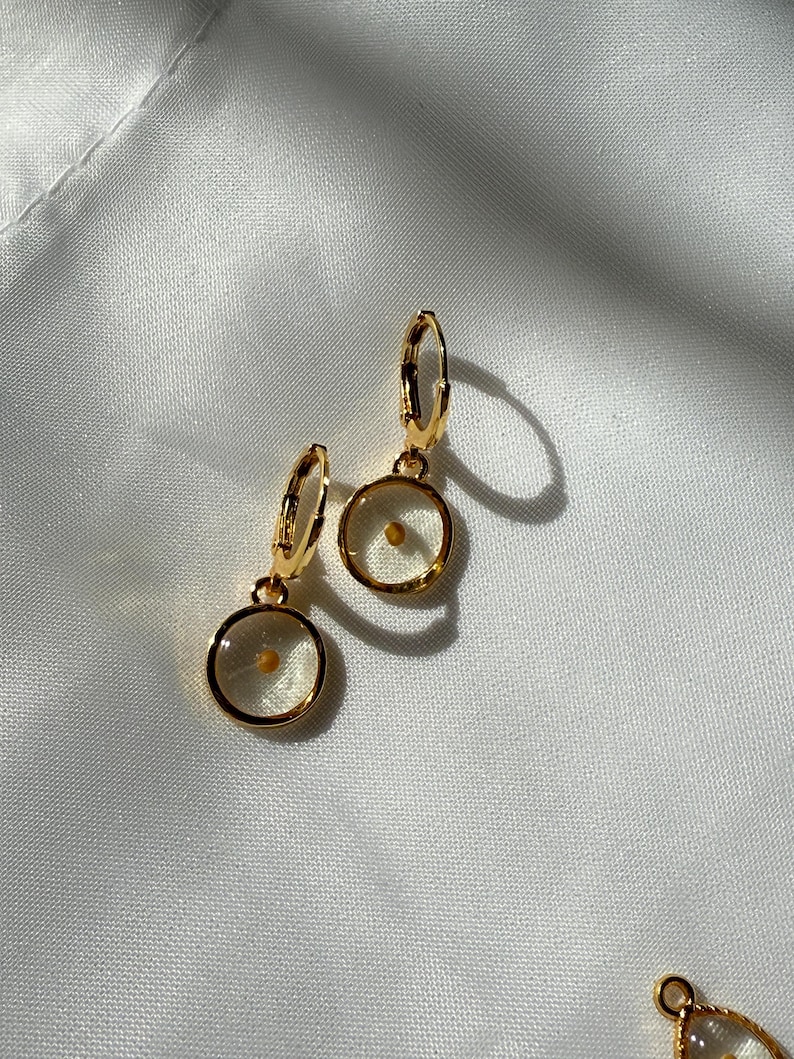May include: A pair of gold hoop earrings with a small, clear circle pendant containing a single seed. The earrings are on a white fabric background.