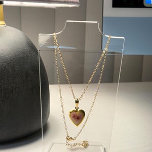 May include: A gold-colored necklace with a heart-shaped pendant. The pendant has a pinkish-red center. The necklace is displayed on a clear acrylic stand. The necklace is a delicate piece of jewelry.