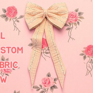 May include: A large, custom fabric bow in a soft peach color with gold trim. The bow is set against a pink background adorned with rose illustrations. Text on the left reads "XXL CUSTOM FABRIC BOW".
