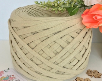 May include: A close-up of a beige t-shirt yarn, a type of yarn made from recycled t-shirts, wound into a large ball. The yarn is woven in a basket weave pattern. The yarn is perfect for making macrame projects.