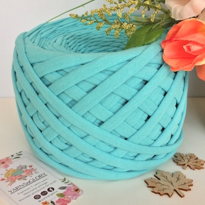 May include: A close-up of a skein of light blue t-shirt yarn. The yarn is woven into a circular shape and is ready to be used for crafting. The yarn is labeled "YarnsGlory" and is described as "Premium T-shirt yarn. Micro-crochet strings. Wooden products for craft."