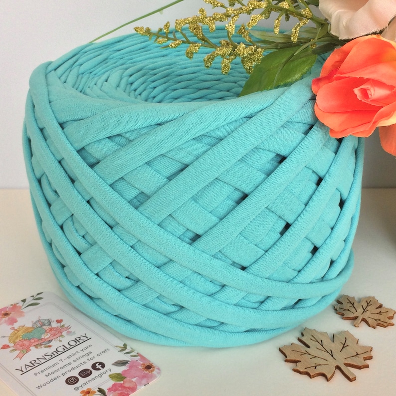 May include: A close-up of a skein of light blue t-shirt yarn. The yarn is woven into a circular shape and is ready to be used for crafting. The yarn is labeled "YarnsGlory" and is described as "Premium T-shirt yarn. Micro-crochet strings. Wooden products for craft."