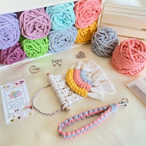 May include: A rainbow macrame wall hanging with a wooden dowel and a gray and pink cord. The macrame is made with cotton rope and has a fringe on the bottom. The wall hanging is surrounded by balls of yarn in various colors, including pink, purple, green, and blue.
