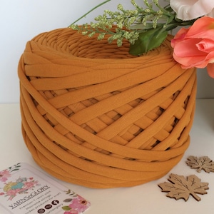 Peut inclure: Une grande bobine de fil de t-shirt jaune moutarde. Le fil est fabriqué à partir de t-shirts recyclés et est parfait pour les projets de macramé. La bobine est posée sur une surface blanche avec une carte de visite qui dit "YarnsGlory" et a une adresse de site web.