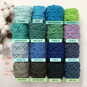 May include: A collection of 16 cotton rope yarn skeins in various shades of blue, green, and purple. The yarn is arranged in four rows of four skeins each. Each skein is labeled with its color name: Tiffany, Aqua, Dark Turquoise, Chartreuse, Caribbean Green, Benet Green, Shamrock, Lavender, Powder Blue, French Blue, Cobalt, Slate Blue, Sage Green, Uniform Green, Emerald, and Dark Teal.