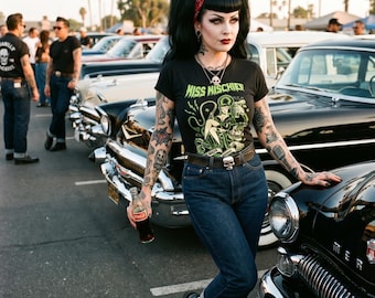 May include: A woman wearing a black t-shirt with "Miss Mischief" and a green graphic, dark blue jeans, and black and white shoes. She holds a Coca-Cola bottle and leans against a classic black car. Other classic cars and people are in the background.