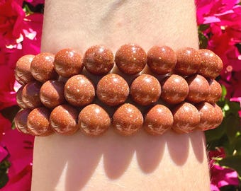 May include: Two bracelets made of round, reddish-brown beads with a glittery appearance. The bracelets are worn on an arm, with a background of pink flowers. Each bead is approximately 1 cm in diameter.