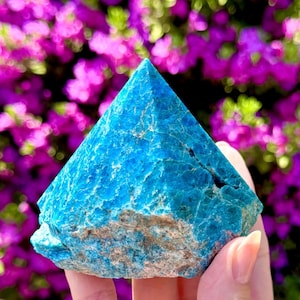 May include: A vibrant blue apatite pyramid gemstone with a textured surface. The gemstone is held in a hand, with a blurred background of purple flowers. The pyramid shape has a pointed top and a rough, natural base.