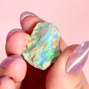 May include: A raw opal gemstone displaying a play of colour with blues, greens, and oranges. The stone has a textured surface and is held against a soft pink background. The opal is a natural, uncut specimen.