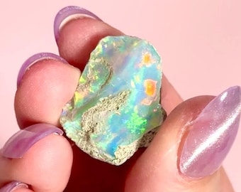 May include: A rough opal gemstone with a vibrant display of colors, including blues, greens, and oranges. The stone has a textured surface and is held against a soft pink background. The opal is a natural, uncut specimen.