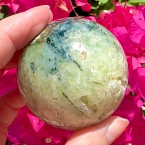 May include: A round, polished gemstone sphere with a mix of green and blue hues. The stone has a textured surface and is held in a person's hand. The background features bright pink flowers.