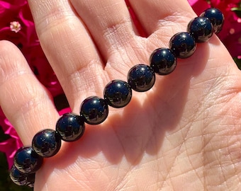 May include: A black beaded bracelet with polished, round beads. The bracelet is held in a hand, with a background of pink flowers.