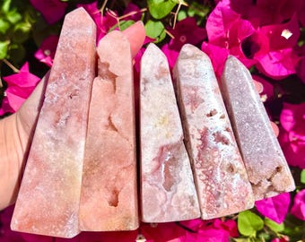 May include: Five pink and white crystal obelisks with varying patterns and textures. The crystals are displayed against a backdrop of bright pink flowers and green foliage, creating a vibrant contrast.