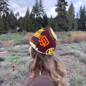 May include: A brown beanie with the San Diego Padres logo in orange and yellow. The beanie is worn by a person with long, wavy brown hair, and a brown fleece jacket. The background features a forest and cloudy sky.