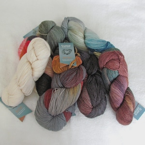 May include: A collection of colorful yarn skeins in various shades of cream, gray, red, orange, and blue. Some skeins have attached tags. The yarn is arranged on a white surface, showcasing the different textures and hues.