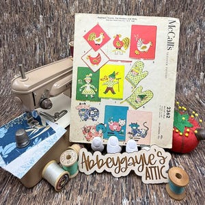 May include: A vintage sewing scene featuring a tan Singer sewing machine, a McCall's transfer pattern, spools of thread, a thimble, and a pin cushion. The pattern includes designs for appliqued towels, pot holders, and mitts with farm animal and folk art motifs.