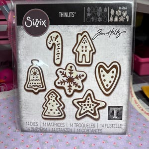 Könnte beinhalten: Eine Packung Sizzix Thinlits Stanzformen mit verschiedenen Weihnachtskeksformen, darunter Zuckerstange, Haus, Glocke, Schneeflocke, Stern, Baum und Herz. Die Stanzformen sind braun mit weißen Details. Die Packung enthält den Text "Sizzix" und "Jim Holtz".