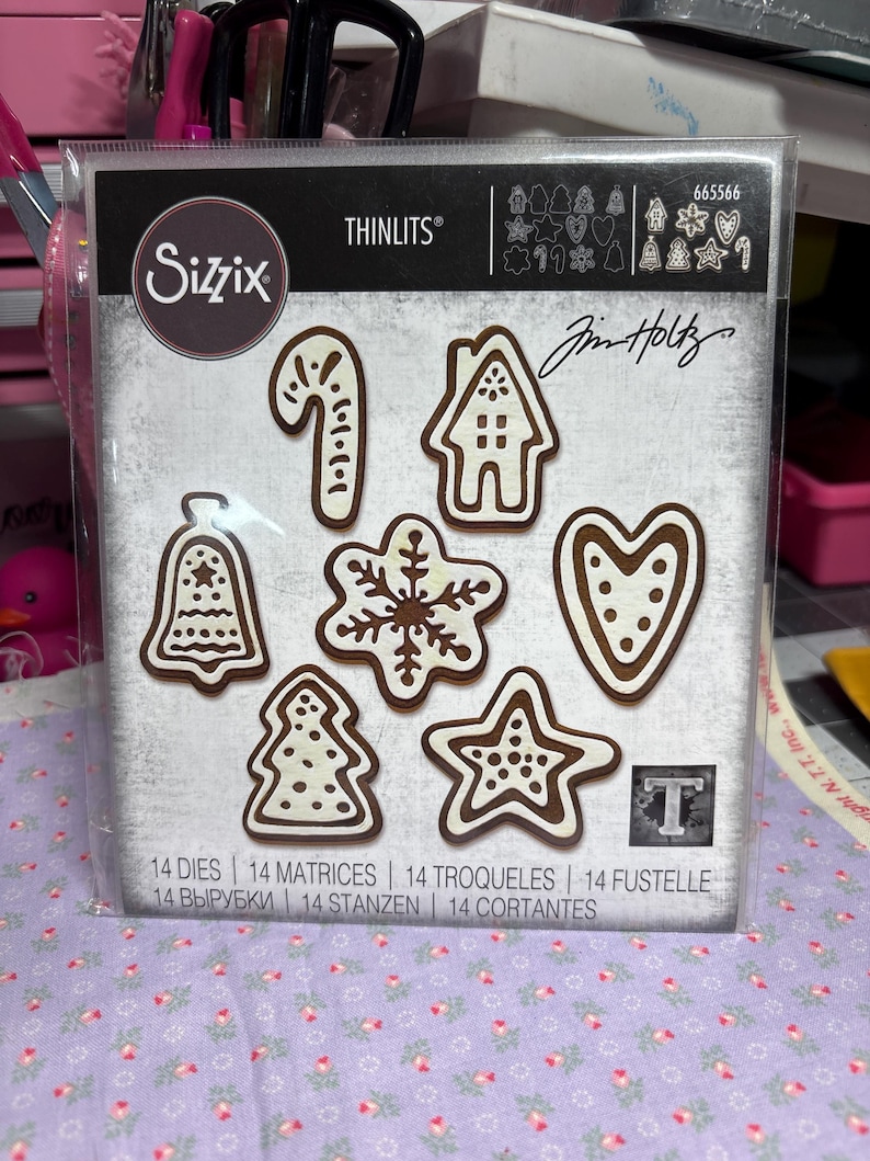 Puede incluir: Un paquete de troqueles Sizzix Thinlits con varias formas de galletas navide&ntilde;as, incluyendo un bast&oacute;n de caramelo, casa, campana, copo de nieve, estrella, &aacute;rbol y coraz&oacute;n. Los troqueles son marrones con detalles blancos. El paquete incluye el texto "Sizzix" y "Jim Holtz".
